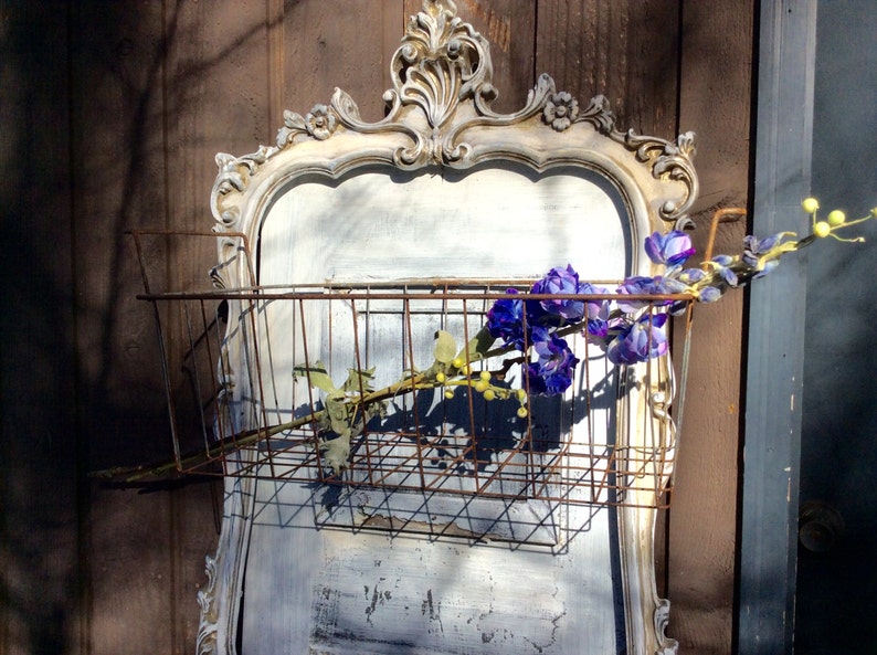 May include: A vintage-style wall-mounted basket with a distressed, ornate frame. The basket is made of rusty metal and holds purple and yellow faux flowers. The frame is painted in a distressed white and gray color scheme.