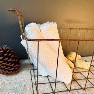 May include: A rustic metal wire basket with a handle, filled with folded white towels. A pine cone and several pieces of natural sponge are visible. The basket has a distressed, aged appearance, suggesting a vintage or farmhouse aesthetic.