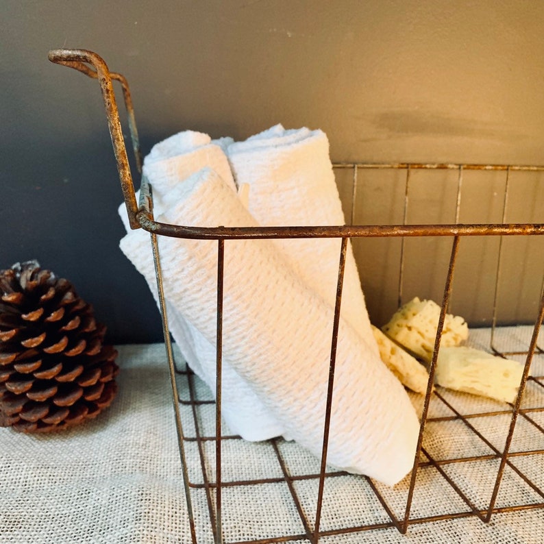 May include: A rustic metal wire basket with a handle, filled with folded white towels. A pine cone and several pieces of natural sponge are visible. The basket has a distressed, aged appearance, suggesting a vintage or farmhouse aesthetic.
