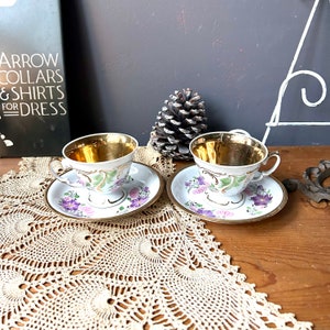 May include: Two white teacups with gold rims and floral designs. The cups are on matching saucers with gold trim and floral designs. The saucers are on a white crocheted doily.