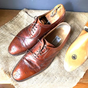 May include: A pair of brown leather wingtip dress shoes with intricate perforations and lace-up closures. The shoes are placed on a burlap surface, with a wooden shoe tree inserted in one shoe. The shoes have a classic, vintage aesthetic.