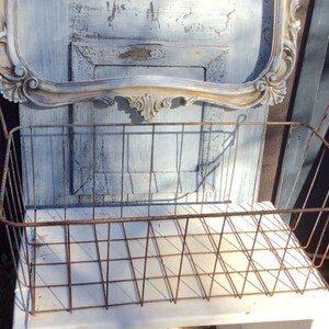 May include: A rustic, rectangular wire basket with a distressed, aged appearance. The basket is made of metal and has a grid pattern. The background includes a vintage-style mirror with ornate detailing and a weathered wooden surface.