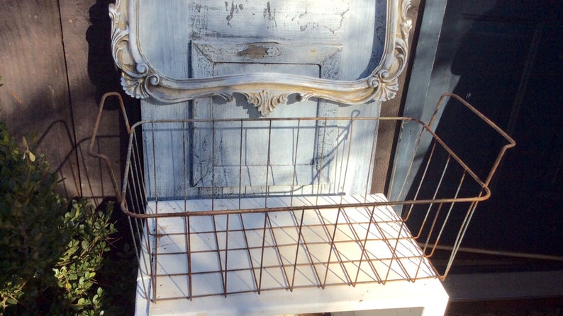 May include: A rustic, rectangular wire basket with a distressed, aged appearance. The basket is made of metal and has a grid pattern. The background includes a vintage-style mirror with ornate detailing and a weathered wooden surface.