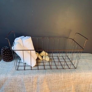 May include: A rectangular, rusty metal wire basket with two handles, holding a rolled white towel and several small, light-colored objects. A pinecone sits to the left of the basket. The basket is on a neutral-colored, textured surface.