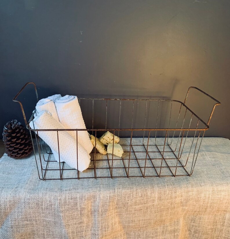 May include: A rectangular, rusty metal wire basket with two handles, holding a rolled white towel and several small, light-colored objects. A pinecone sits to the left of the basket. The basket is on a neutral-colored, textured surface.