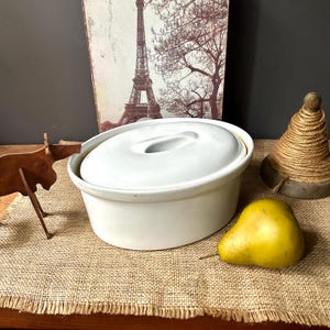 May include: A white ceramic casserole dish with a matching lid. The dish is round with a handle on the lid. A yellow pear sits next to the dish. A decorative Eiffel Tower print is in the background.
