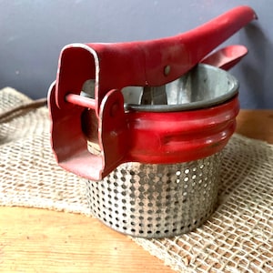 May include: Red metal potato masher with a perforated metal bowl. The masher has a handle with a lever to press down on the potatoes.