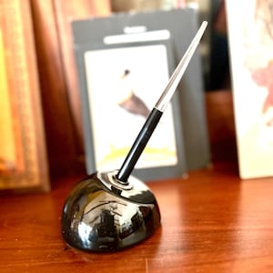 May include: A black ink pen holder with a shiny, rounded base. The pen has a black shaft and a clear, pointed tip. The pen holder is sitting on a wooden surface.