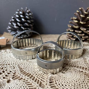 May include: Three vintage, silver-toned cookie cutters with handles. The cutters are round with fluted edges. The cutters are arranged on a cream-colored, crocheted doily, with pine cones in the background.