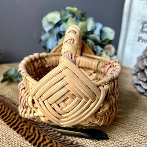 May include: A small, woven basket with a handle. The basket is made of natural materials and has a light brown color. The basket is sitting on a brown, textured surface.
