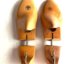 May include: Two wooden shoe trees with a light brown finish. Each tree has a rounded toe and a heel section connected by a metal rod. A circular logo is visible on the top of each tree.