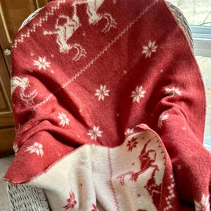 May include: A red and white patterned blanket draped over a wicker chair. The blanket features a repeating pattern of white reindeer and snowflakes on a red background. The underside is white with a red pattern.