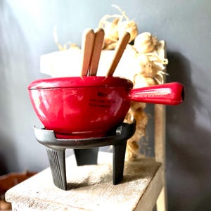 May include: A bright red enameled cast iron fondue pot with a single side handle, resting on a black metal stand. Three light-colored wooden fondue forks are placed inside the pot. The pot features a glossy finish and a visible black text logo.
