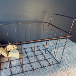 May include: A rectangular, rusty metal wire basket with a grid pattern and two handles. The basket contains several pieces of light-colored, textured material. The basket sits on a light blue fabric, against a dark gray background.