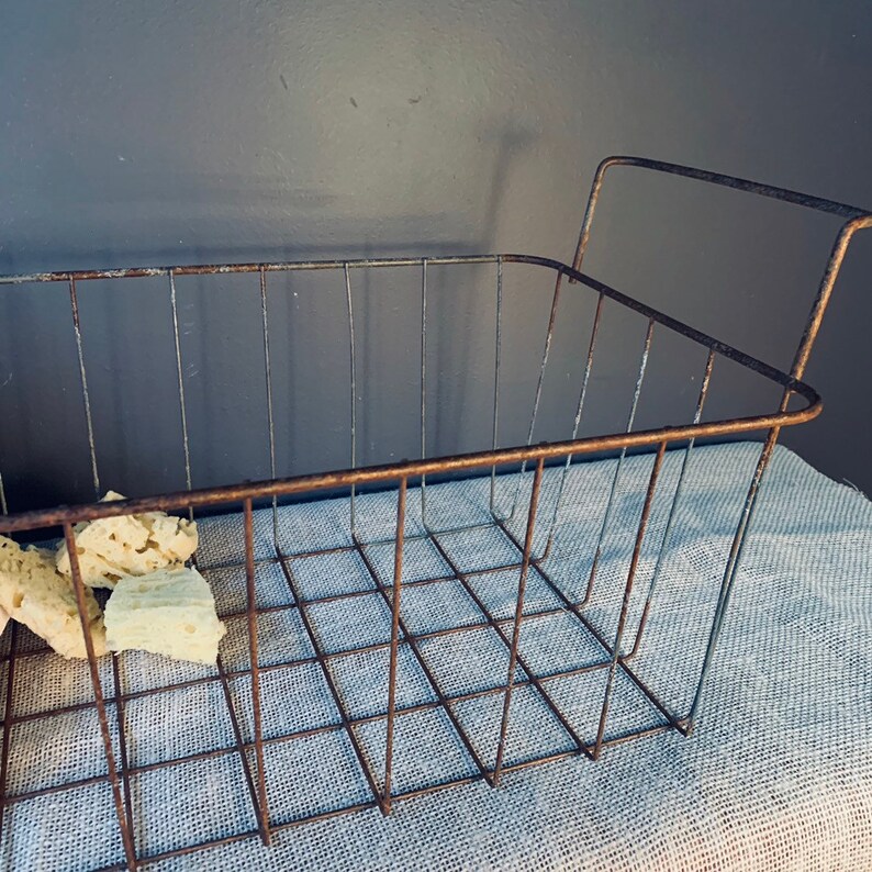 May include: A rectangular, rusty metal wire basket with a grid pattern and two handles. The basket contains several pieces of light-colored, textured material. The basket sits on a light blue fabric, against a dark gray background.