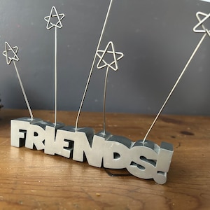 May include: Silver metal 'FRIENDS' sign with five star-shaped wire photo holders.