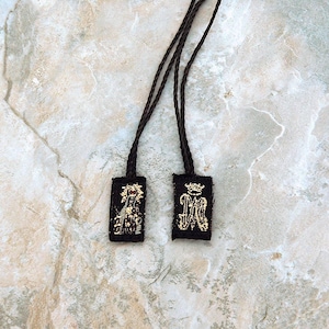 May include: Two black scapular necklaces with woven designs. One features a religious figure with a red halo, the other displays a gold-colored crown and the letter "M". Each is suspended from a brown cord.