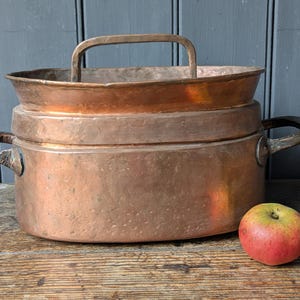 May include: A large, antique copper cooking pot with a handle and two side handles. The pot is sitting on a wooden surface next to a red and yellow apple. The pot has a round base and a flared rim.