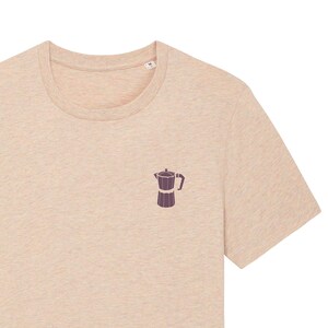 Könnte beinhalten: Ein beiges T-Shirt mit einer kleinen, dunkelvioletten Illustration einer Kaffeekanne auf der Vorderseite.