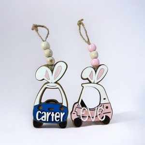May include: Two wooden bunny-shaped car ornaments with the names "carter" and "eve" written on them. The car with the name "carter" is blue and the car with the name "eve" is pink.