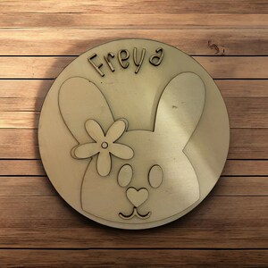 May include: A round wooden plaque with a bunny rabbit design. The bunny has a flower on its head and the name "Freyja" is written above it.