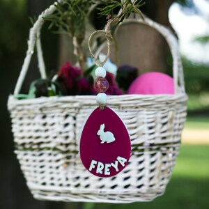May include: A pink Easter egg ornament with a white bunny silhouette and the name "FREYA" written on it. The ornament is hanging from a white wicker basket filled with flowers.