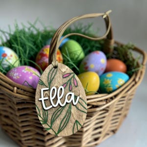 May include: A wooden Easter egg tag with the name "Ella" in white script, adorned with green leaf and pink floral accents. The tag hangs over a wicker basket filled with colorful, decorated Easter eggs and green decorative grass.