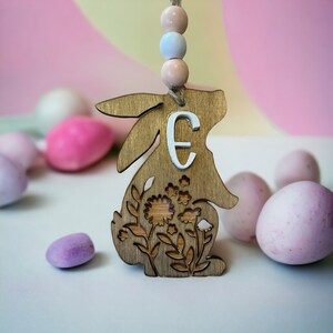 May include: Wooden bunny cutout with a floral design and the letter "E" painted on it. The bunny is hanging from a string of pastel beads. The bunny is surrounded by pastel pink Easter eggs.