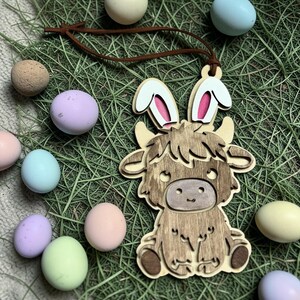 May include: Wooden Highland cow ornament with bunny ears, brown leather cord, and colorful Easter eggs. The cow is light brown with a darker brown face and fur details. Eggs are pastel shades of pink, blue, green, and yellow.
