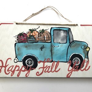 May include: A rectangular wooden sign with a painted illustration of a vintage blue pickup truck carrying pumpkins. The sign has the text "Happy Fall Y'all" in red script. A twine loop is attached for hanging.