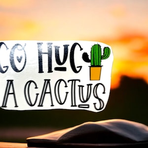 May include: A white sticker with black text that reads "So  Hug  A Cactus" with a small green cactus in a yellow pot. The sticker is on a wooden surface with a blurred sunset background.