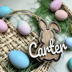 May include: Wooden Easter bunny-shaped ornament with the name "Carter" in white script. The ornament is brown with a dark blue outline and a light brown string. Pastel-colored Easter eggs surround the ornament in a woven basket.