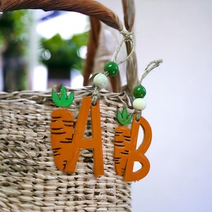 May include: A wicker basket with a wooden carrot-shaped letter A and B hanging from it. The letters are orange with green carrot tops. The basket is decorated with green and white beads.