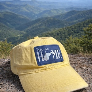 May include: Yellow baseball cap with a blue patch displaying the text "Take me HOME" and a white outline of West Virginia. The cap is set against a scenic mountain backdrop.