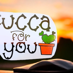 May include: White sticker with black text that reads "Succa for you" and a cartoon cactus in a red pot. The sticker is on a black surface against a sunset background.
