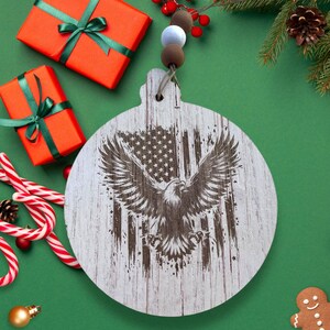 May include: White wooden Christmas ornament with a distressed finish. The ornament features a brown eagle with wings spread wide, flying in front of an American flag.