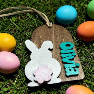 May include: Wooden Easter tag with a white bunny silhouette and the name "Olivia" in teal. The tag is brown with a leather string for hanging. Colorful Easter eggs in yellow, pink, blue, and orange surround the tag.