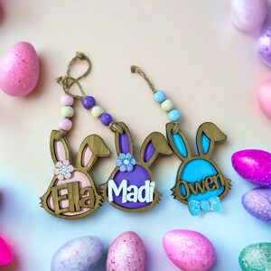 May include: Three wooden bunny ornaments with names "Ella", "Madi", and "Owen" written on them. Each bunny is decorated with a flower and has a string of colorful beads attached.