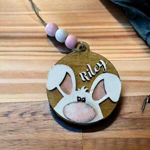 May include: A wooden Easter bunny ornament with a pink nose, white ears, and black eyes. The ornament is painted with the name "Riley" in white lettering.