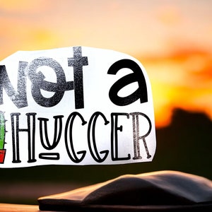 May include: White sticker with black text that reads "Not a hugger" with a small cactus illustration in a red pot. The sticker is against a blurred background of an orange sunset.