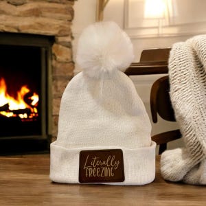 May include: A white knit beanie with a fluffy pom-pom on top. The beanie has a brown leather patch with the words "Literally Freezing" in a script font. A cozy winter accessory.