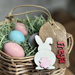 May include: A wicker basket filled with faux green grass and pastel-colored Easter eggs. A wooden tag with the name "Freya" in red letters and a white bunny cutout with a pink flower tail is attached to the basket.