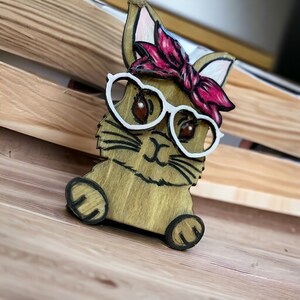 May include: A wooden cutout of a bunny wearing heart-shaped glasses and a pink bandana. The bunny has brown fur and white whiskers.