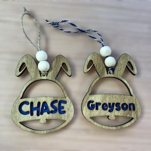 May include: Two wooden bunny-shaped ornaments with white beads and blue painted names, "CHASE" and "GREYSON", on a light brown wooden surface.