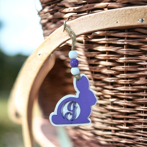 May include: A purple wooden bunny cutout with a white circle in the center that has the letter "J" in it. The bunny is hanging from a wicker basket with a string of purple and white beads.