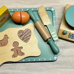 May include: Wooden toy kitchen set with a rolling pin, knife, and cutting board with shapes. Includes a scale with a digital display, a yellow block, and an orange egg. The set is painted in shades of green, brown, and yellow.