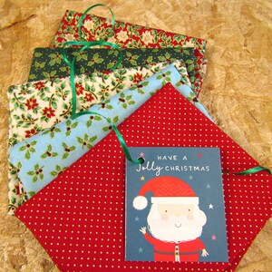 K&ouml;nnte beinhalten: Ein Stapel aus f&uuml;nf Stoffquadraten mit einem weihnachtlichen Druck. Die Stoffquadrate sind in Rot, Gr&uuml;n, Blau und Wei&szlig; mit einem Muster aus Stechpalmenbl&auml;ttern und Beeren. Ein Geschenkanh&auml;nger mit dem Text "Have a Jolly Christmas" und einem Comic-Weihnachtsmann ist an den Stoffquadraten befestigt.