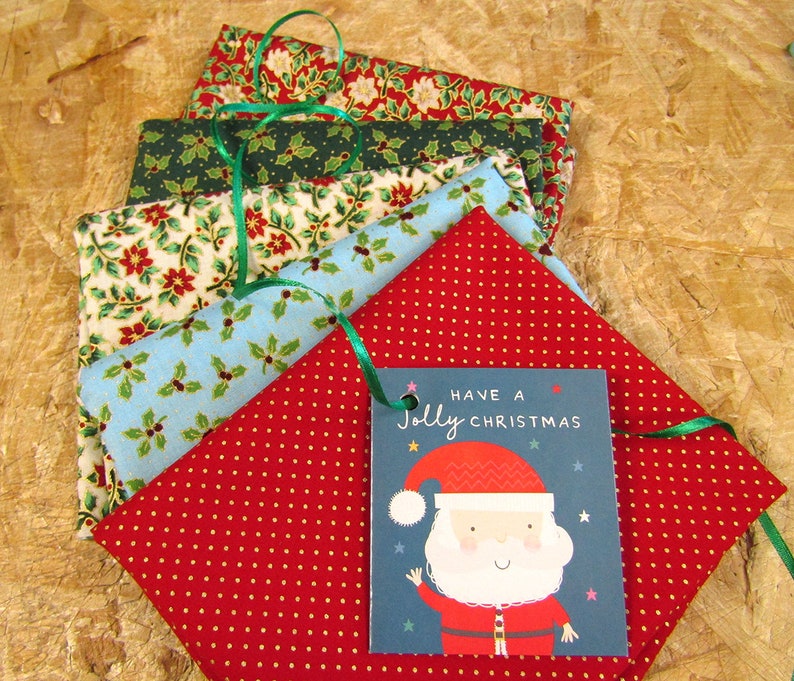 K&ouml;nnte beinhalten: Ein Stapel aus f&uuml;nf Stoffquadraten mit einem weihnachtlichen Druck. Die Stoffquadrate sind in Rot, Gr&uuml;n, Blau und Wei&szlig; mit einem Muster aus Stechpalmenbl&auml;ttern und Beeren. Ein Geschenkanh&auml;nger mit dem Text "Have a Jolly Christmas" und einem Comic-Weihnachtsmann ist an den Stoffquadraten befestigt.