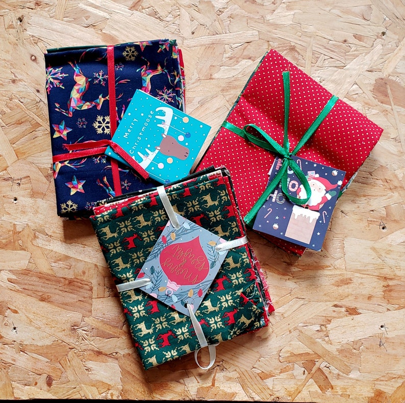 K&ouml;nnte beinhalten: Ein Set aus vier Weihnachtsstoffquadraten, die mit rotem und gr&uuml;nem Band umwickelt sind. Die Stoffquadrate zeigen verschiedene festliche Designs, darunter Rentiere, Schneeflocken und einen Elch. Die Quadrate sind mit einem roten Band und einer gr&uuml;nen Schleife zusammengebunden. Die Stoffquadrate sind perfekt zum Basteln, Quilten oder Dekorieren.