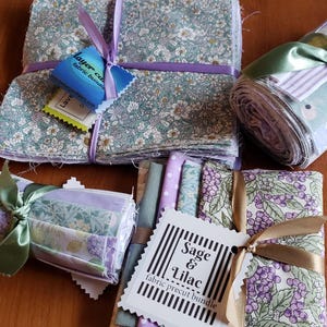 Puede incluir: Paquetes de tela con diseños florales y estampados, atados con cintas. Los paquetes incluyen cuadrados y tela enrollada, con etiquetas adjuntas. Los colores incluyen verde salvia, lavanda y blanco.